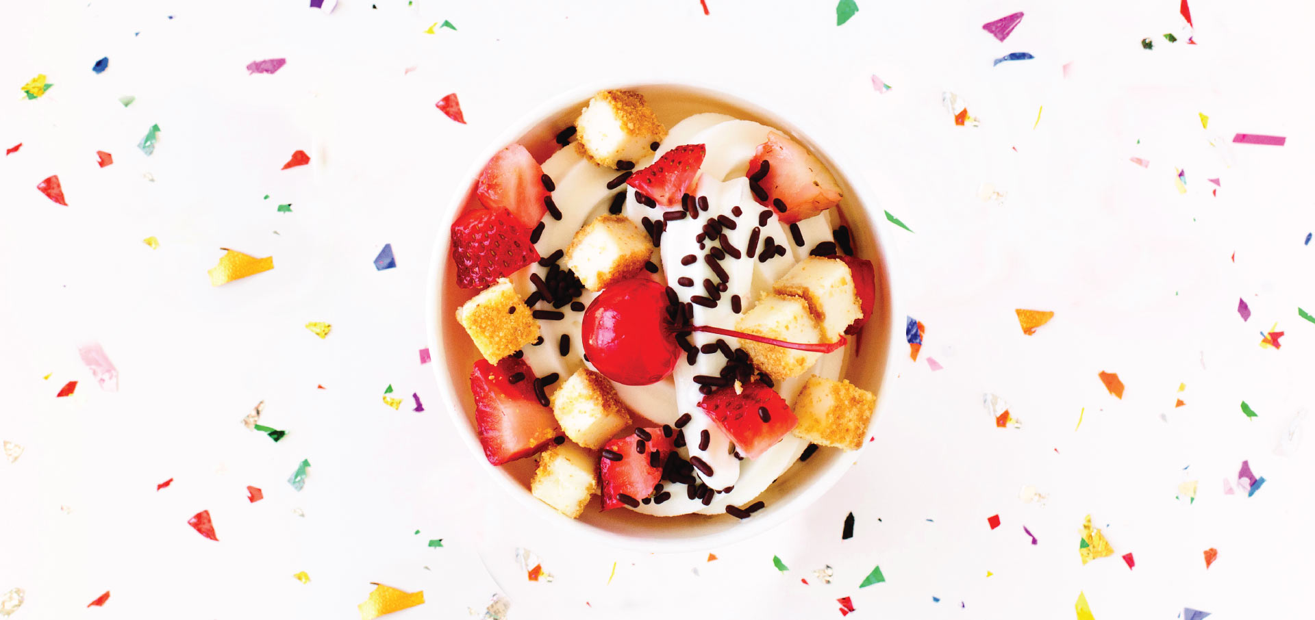 A bowl viewed from the top with fresh fruit, frozen yogurt, and chocolate sprinkles and a background of confetti on white